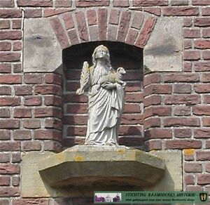 Heilige Agnes Raamsdonk – Kerkplein 3, aan noordzijde van parkeerplaats tussen kerk en Ontmoetingscentrum 1937 – In een nis boven de ingang van de voormalige katholieke bewaarschool staat het beeld van de heilige Agnes. De naam Agnes komt van het Griekse hagnès,wat de kuise betekent. De heilige Agnes draagt in haar linkerarm een lam en in haar rechterhand een palmtak. De palmtak is het christelijke symbool voor de overwinning op de dood door en in Christus. Zo worden martelaren vaak met een palmtak afgebeeld. Het lam verwijst naar het Latijnse woord agnus, dat lam betekent en symbool staat voor onschuld en zuiverheid. Het hardstenen beeld is in 1937 gemaakt door de Raamsdonker Jan Kappe, timmerman, beeldhouwer en schrijnwerker. Agnes is de beschermheilige van maagden, verliefden dan wel verloofden en tuinlieden. De achtergrond hiervan ligt in haar eigen levensverhaal. De mooie Agnes leefde in de derde eeuw na Christus in Rome en werd al op jonge leeftijd ten huwelijk gevraagd door de zoon van de stadsprefect. Ondanks de martelingen die ze moest ondergaan, weigerde Agnes omdat zij voor zichzelf een toekomst zag als bruid van Christus (non). Uiteindelijk werd ze gedood. Na haar dood zou ze aan haar ouders zijn verschenen met een lam (de onschuld) aan haar zijde. In 1937 breidden de Zusters van Liefde het Adrianusgebouw uit met kleuterschool St. Agnes.
